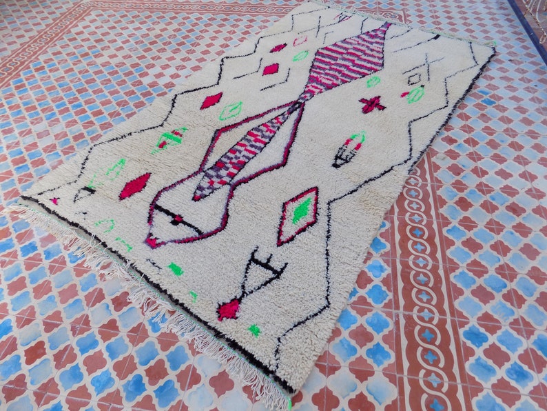 May include: A white Moroccan rug with a geometric pattern in shades of pink, red, green, and black. The rug has a fringe border and is laid out on a tiled floor.