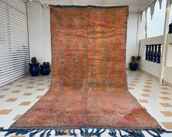 May include: A vintage Moroccan rug with a geometric pattern in shades of brown, red, and blue. The rug has a fringe border and is laid out on a tiled floor.