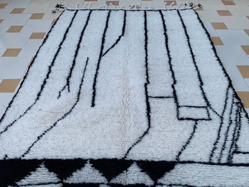 May include: A white and black Moroccan rug with a geometric pattern. The rug has a fringe border and is made of wool.