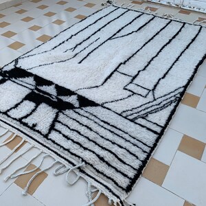 May include: A white and black geometric patterned rug with fringe on the edges. The rug is laid out on a tiled floor.