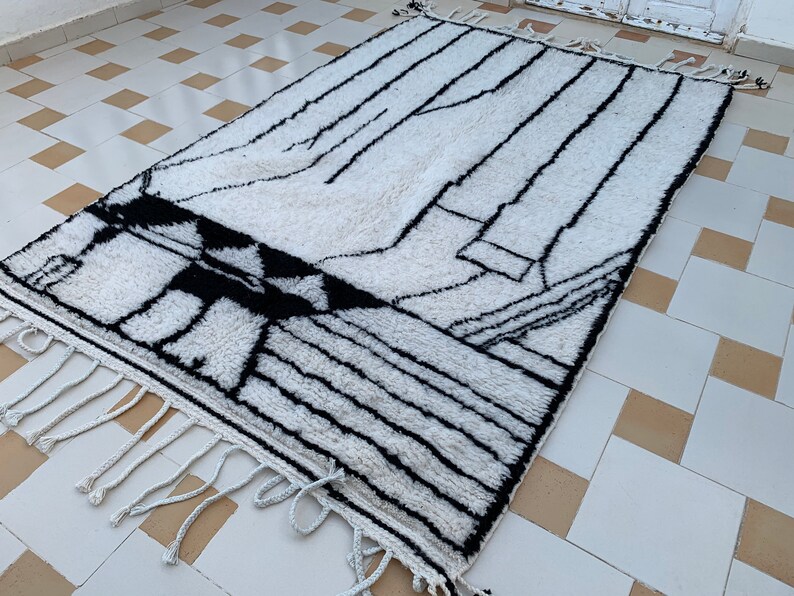 May include: A white and black geometric patterned rug with fringe on the edges. The rug is laid out on a tiled floor.