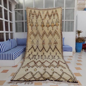 May include: A rectangular, cream-colored rug with a geometric pattern in brown and tan. The rug features a series of zigzags, diamonds, and other shapes. The rug is displayed vertically against a white wall, with a blue and white striped bench in the background.
