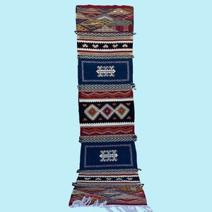 May include: A long, narrow rug with a colorful geometric pattern. The rug is made up of several panels, each with a different design. The colors include red, blue, white, black, and brown.