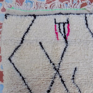 May include: White wool rug with a black and pink geometric pattern. The rug has a fringe border.