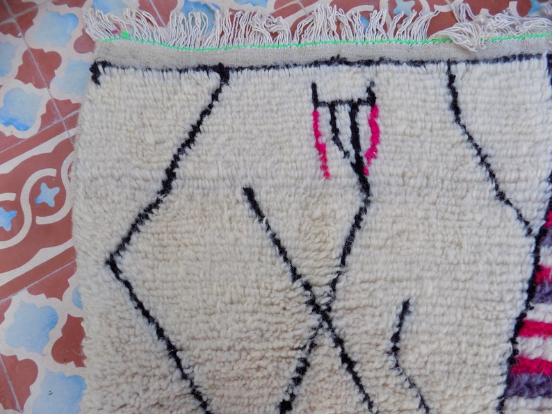 May include: White wool rug with a black and pink geometric pattern. The rug has a fringe border.