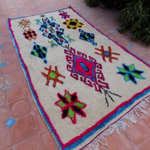 May include: A white Moroccan rug with a colorful geometric pattern. The rug has a fringe border and features a variety of shapes and colors, including pink, blue, green, and orange.