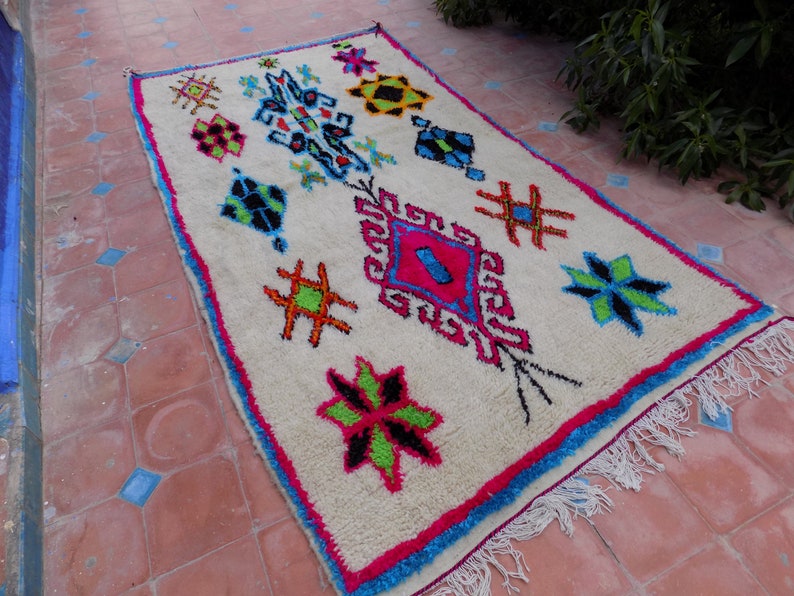 May include: A white Moroccan rug with a colorful geometric pattern. The rug has a fringe border and features a variety of shapes and colors, including pink, blue, green, and orange.
