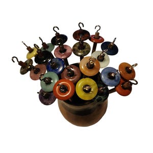May include: A collection of drop spindles featuring circular whorls made from various colorful stones and minerals, each attached to a wooden shaft with a metal hook at the top. The spindles are arranged upright in a dark, rounded wooden container.
