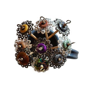 May include: A collection of drop spindles featuring wooden shafts and decorative filigree metal whorls in various colors including purple, brown, green, and black, arranged in a dark mug. Each spindle is topped with a small metal hook.