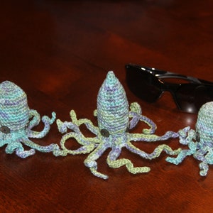 Könnte beinhalten: Drei gehäkelte Octopus-Plüschtiere in verschiedenen Größen, aus grünem, blauem und violettem Garn. Die Plüschtiere sind auf einer Holzoberfläche angeordnet.