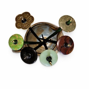 May include: An overhead shot of a decorative arrangement featuring a ceramic bowl surrounded by various stone and metal accents. The arrangement includes green, black, and brown circular stones, a gold-colored flower-shaped piece, and dark green stems.