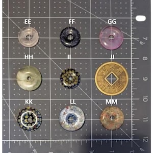 May include: A collection of nine circular beads with center holes arranged in a grid on a cutting mat with inch markings. The beads include translucent pink, dark blue, light green, floral millefiori patterns, and a gold-toned coin-shaped piece.