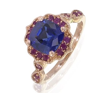 May include: A gold ring featuring a large, square, deep blue gemstone surrounded by smaller, round, red gemstones. The ring has a decorative, floral-inspired setting and a delicate band, showcasing a vintage or antique style.