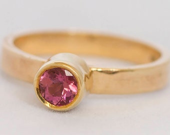 Puede incluir: Un anillo de oro con una piedra preciosa rosa engastada en un engaste de bisel. La banda del anillo es texturizada y tiene un acabado martillado.