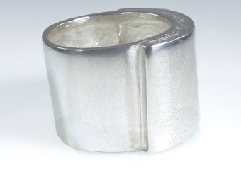 May include: A silver ring with a wide band and a flat, rectangular top. The ring has a smooth, polished finish.