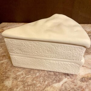 Op de afbeelding: Een witte keramische cakebox in de vorm van een stuk cake. De doos heeft een getextureerd oppervlak en een verhoogde rand aan de bovenkant.