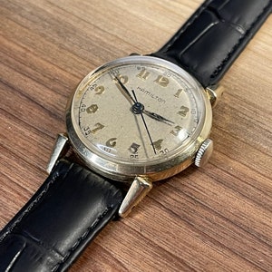 May include: A gold-toned wristwatch with a white face and black hands. The watch has black Roman numeral hour markers and a black leather strap. The watch face says "Hamilton".