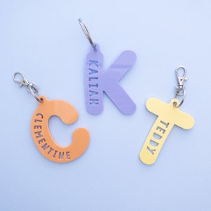 Puede incluir: Tres llaveros personalizados de colores con los nombres "Kaliah", "Clementine" y "Teddy" recortados en acrílico. Los llaveros tienen la forma de la primera letra de cada nombre.