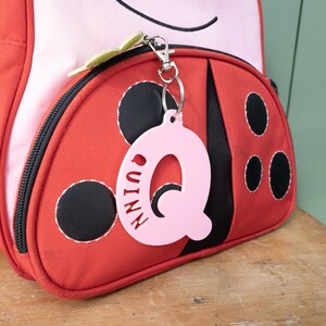 May include: Pink plastic letter Q keychain with the name "Quinn" written on it. The keychain is attached to a red ladybug backpack.