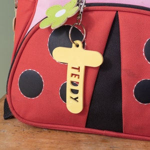 May include: A red ladybug-themed backpack with black polka dots and a yellow wooden name tag that says "TEDDY" hanging from a silver key chain.