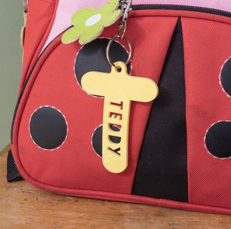 May include: A red ladybug-themed backpack with black polka dots and a yellow wooden name tag that says "TEDDY" hanging from a silver key chain.