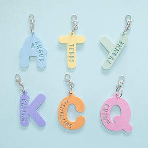 May include: Six colorful keychains with different letters and names, including Angus, Teddy, Yvonne, Kaliah, Clementine, and Quinn. The keychains are made of acrylic and have a silver metal ring.