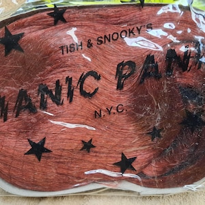 May include: A package of red hair extensions with the text "MANIC PANIC N.Y.C." and "TISH & SNOOKY'S" printed on the front. Black star shapes are scattered across the front of the package.