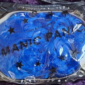 May include: A blue wig in a clear plastic bag. The bag has black text that reads "MANIC PANIC NYC" and "TISH & SNOOKY'S". Black star shapes are scattered across the wig. The wig is a vibrant shade of blue.