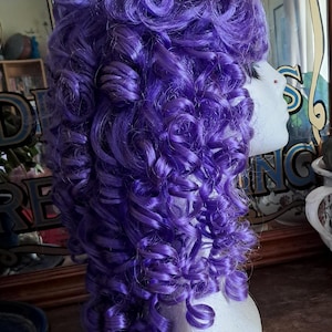 May include: A vibrant purple wig with voluminous, tightly curled ringlets. The wig is styled on a white foam head, showcasing the intricate curls and rich color. The curls cascade down, creating a full and textured look, perfect for cosplay or costume events.