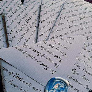 May include: A collection of light blue envelopes with handwritten text about Tarot cards. A blue and white wax seal is visible on one envelope. The envelopes are arranged in a fan shape, with the text and seal in focus.