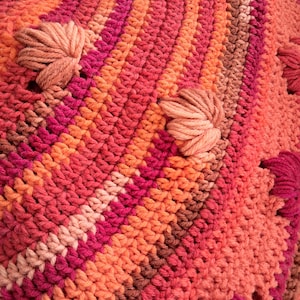 May include: A crocheted textile featuring a vibrant gradient of warm colours including shades of coral, orange, magenta, and brown. The surface displays a repeating pattern of textured yarn clusters interspersed between rows of dense, uniform crochet stitches.