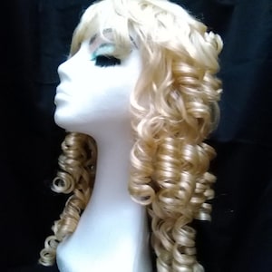 May include: A blonde wig with long, tight curls is displayed on a white mannequin head. The wig has a side part and is styled with voluminous ringlets. The background is a solid black.
