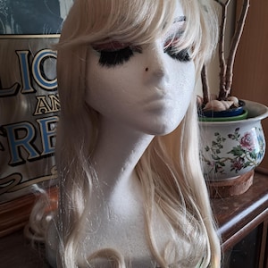 May include: A blonde wig with long, wavy hair and bangs is displayed on a white mannequin head. The wig has a natural-looking style and color. The mannequin head has painted eyes and eyelashes. The wig is likely for cosplay or costume use.