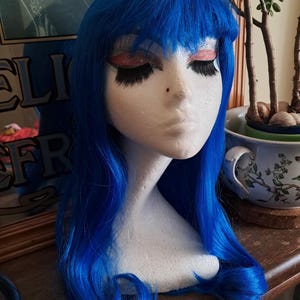 May include: A blue wig with bangs and long, wavy hair is displayed on a white mannequin head. The mannequin has long, black eyelashes and pink eyeshadow. The wig is a vibrant shade of blue, and the overall effect is striking.