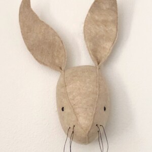 May include: A beige felt bunny head with long ears and black thread whiskers. The bunny head is a wall hanging decoration.