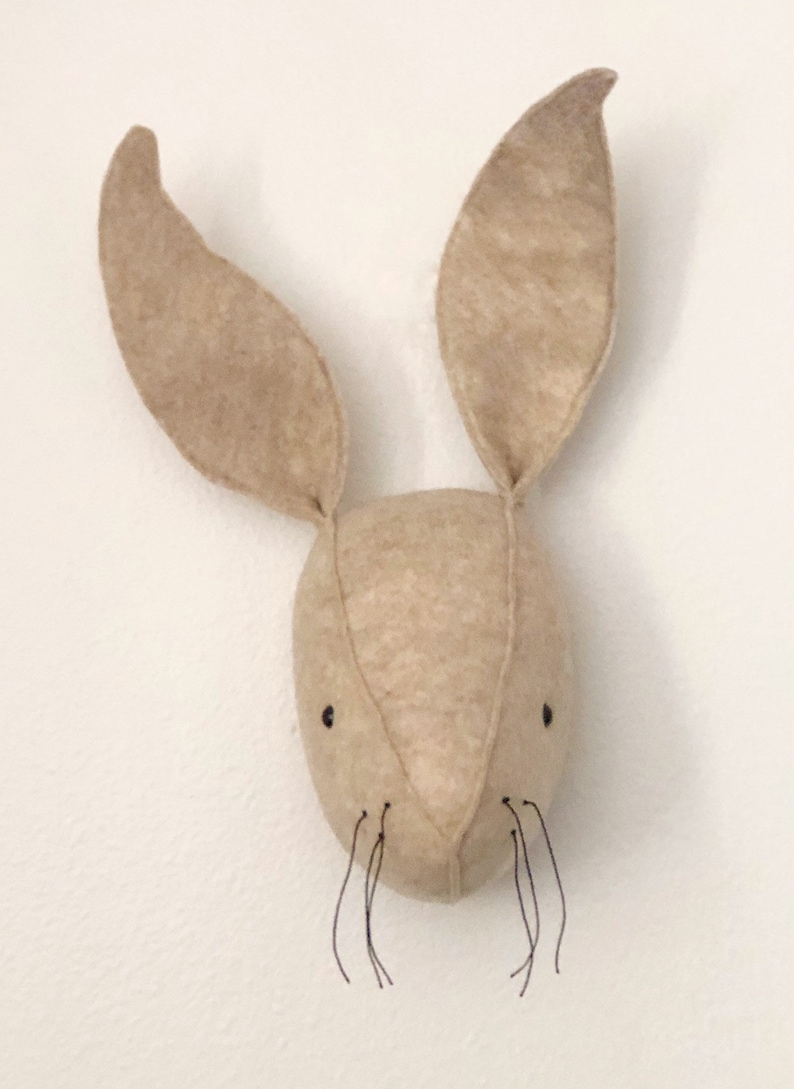 May include: A beige felt bunny head with long ears and black thread whiskers. The bunny head is a wall hanging decoration.