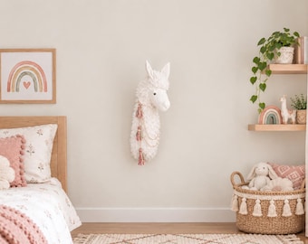 May include: A white plush llama wall mount featuring a textured, shaggy coat and decorative hanging tassels in muted pink and cream tones. The llama head is mounted on a plain wall, positioned between a bed with pink bedding and wooden shelves.