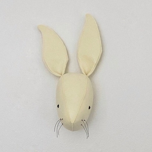 May include: A cream-colored felt bunny head with black eyes and whiskers. The bunny head is a wall decoration.