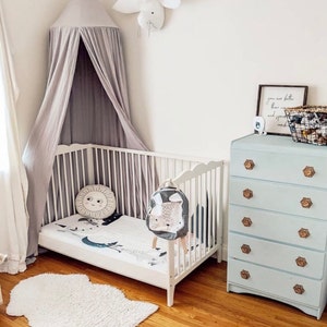 May include: A white crib with a grey canopy and a white rug. A light blue dresser with gold knobs is in the background. The crib has a white and grey bedding set with a sun and moon design. A backpack with a bunny face is hanging on the crib.