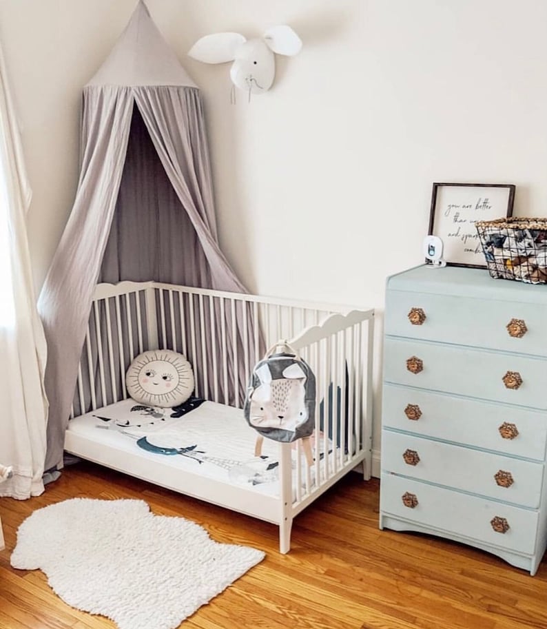 May include: A white crib with a grey canopy and a white rug. A light blue dresser with gold knobs is in the background. The crib has a white and grey bedding set with a sun and moon design. A backpack with a bunny face is hanging on the crib.