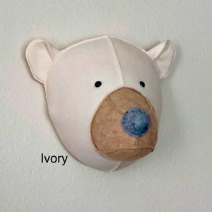 May include: A white felt bear head wall decor with a brown felt nose and blue felt eyes. The bear is mounted on a white wall. The text "Ivory" is visible below the bear.