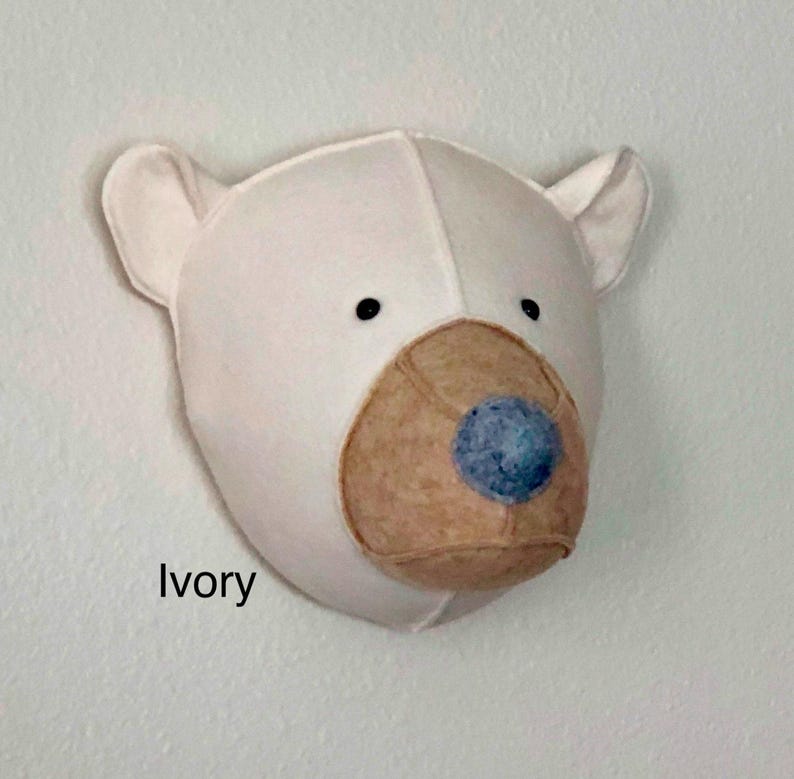 May include: A white felt bear head wall decor with a brown felt nose and blue felt eyes. The bear is mounted on a white wall. The text "Ivory" is visible below the bear.