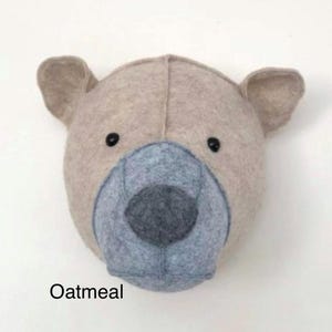 May include: A beige felt animal head wall decor in the shape of a bear with a gray felt nose and black eyes. The bear is labeled "Oatmeal".