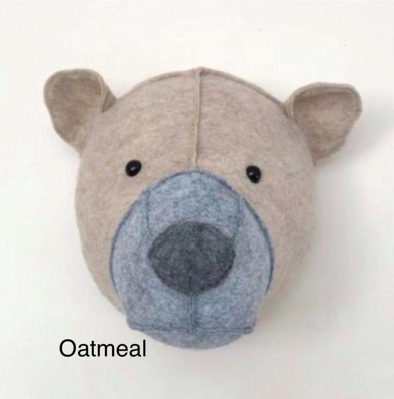 May include: A beige felt animal head wall decor in the shape of a bear with a gray felt nose and black eyes. The bear is labeled "Oatmeal".
