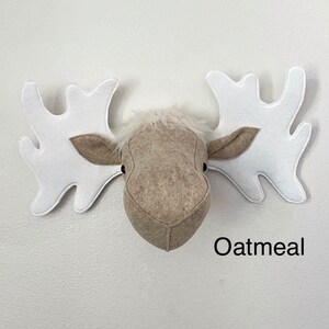 May include: A beige felt moose head with white felt antlers. The moose head is a wall decoration and has the word "Oatmeal" written below it.