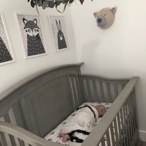May include: A gray crib with a baby sleeping inside. The crib has a white and black patterned sheet. There are three black and white animal prints on the wall above the crib. A stuffed animal bear head is on the wall to the right of the crib.