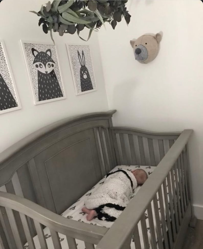 May include: A gray crib with a baby sleeping inside. The crib has a white and black patterned sheet. There are three black and white animal prints on the wall above the crib. A stuffed animal bear head is on the wall to the right of the crib.
