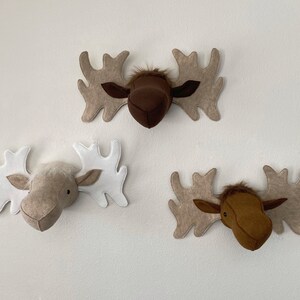 May include: Three felt moose head wall decorations. One is brown with beige antlers, one is beige with white antlers, and one is brown with beige antlers.