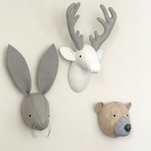 May include: Three felt animal head wall decorations. A white deer head with gray antlers, a gray rabbit head with long ears, and a brown bear head with gray fur.