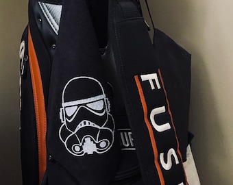 May include: Black golf bag with an orange stripe and a white "FUSION" logo on the strap. A black and white Stormtrooper graphic is on a black towel hanging from the bag.